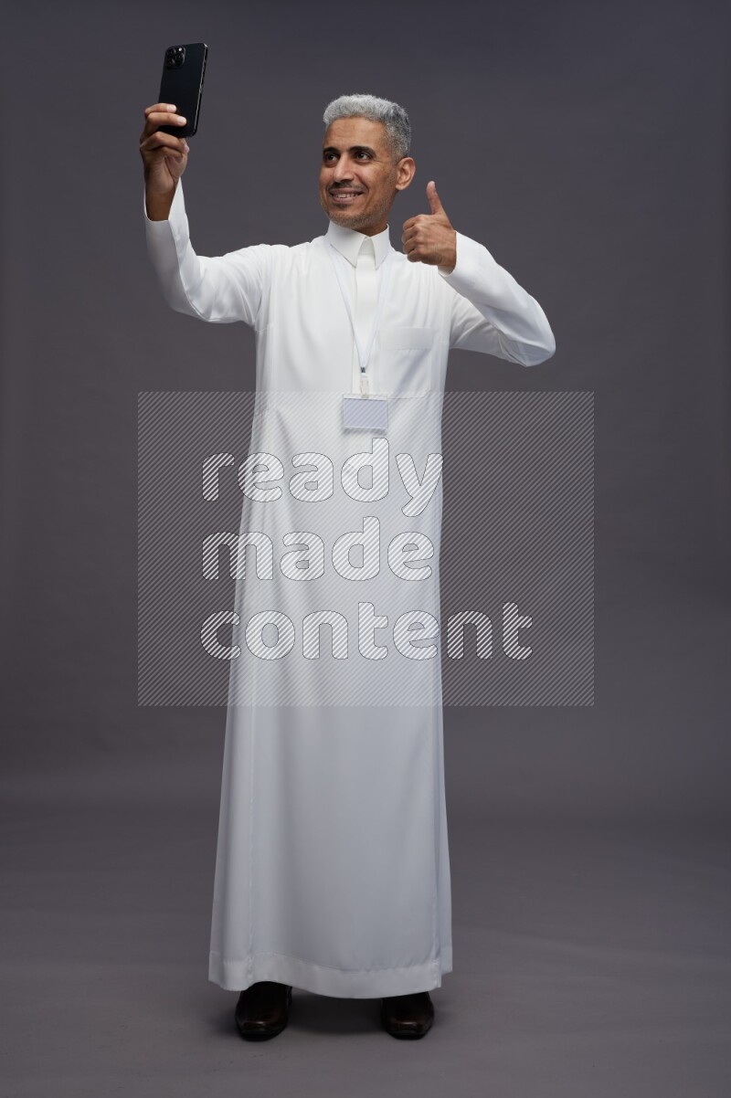 Saudi man wearing thob with neck strap employee badge standing taking selife on gray background