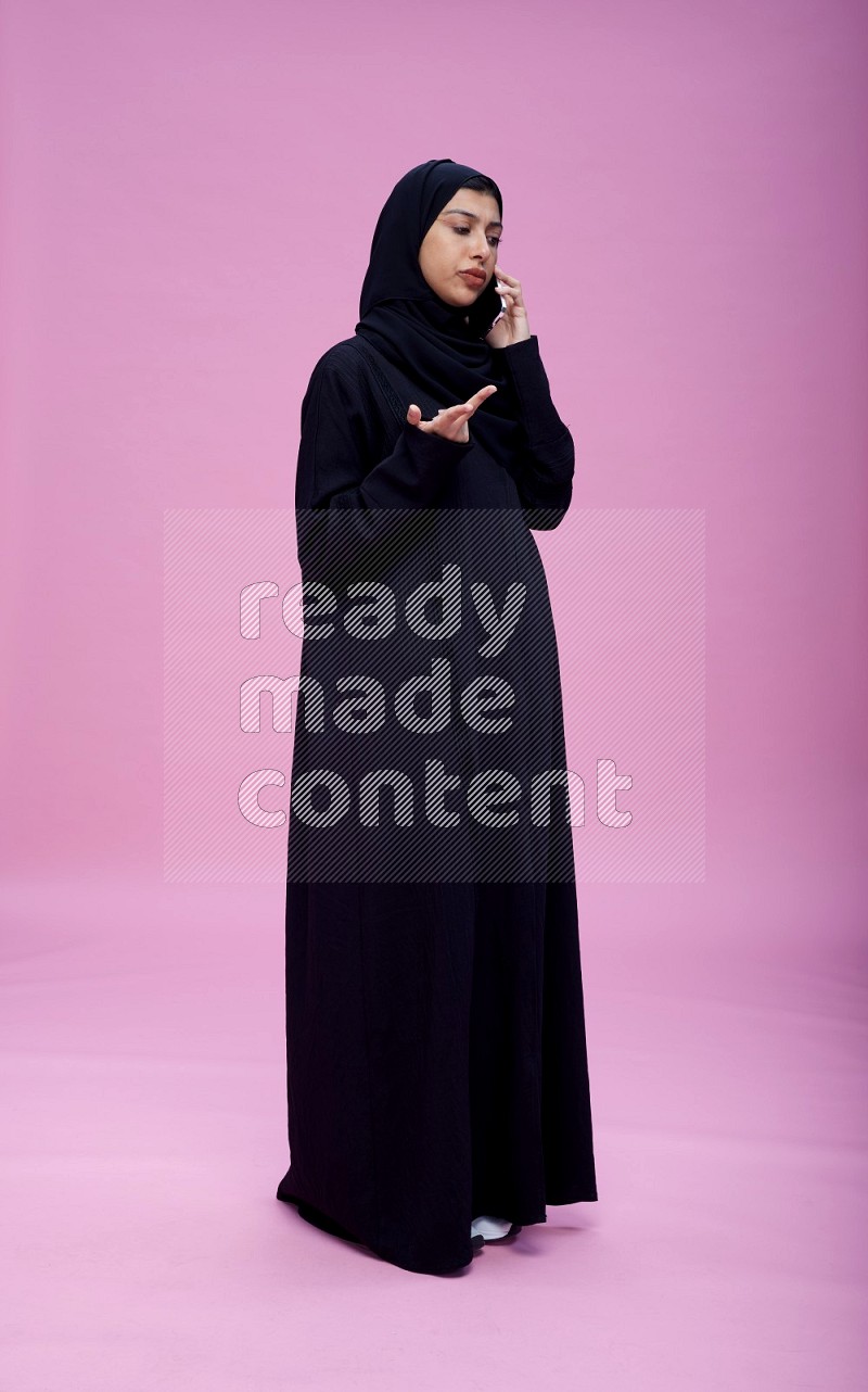 Saudi woman wearing Abaya standing talking on phone on pink background