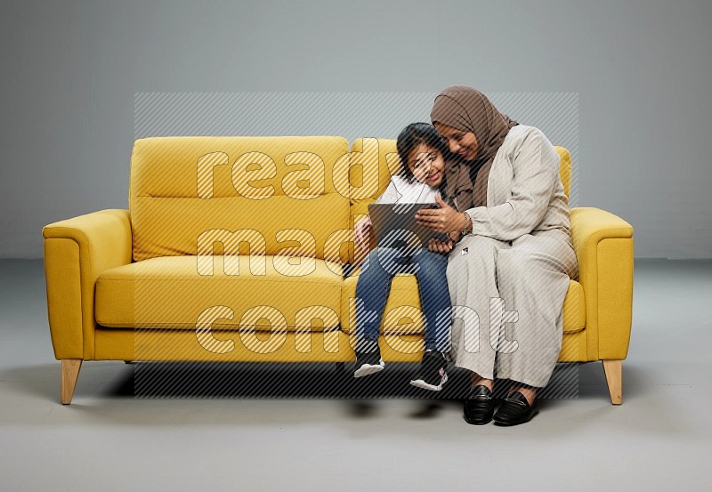 A girl with her mother on yellow sofa and watching on iPad on gray background