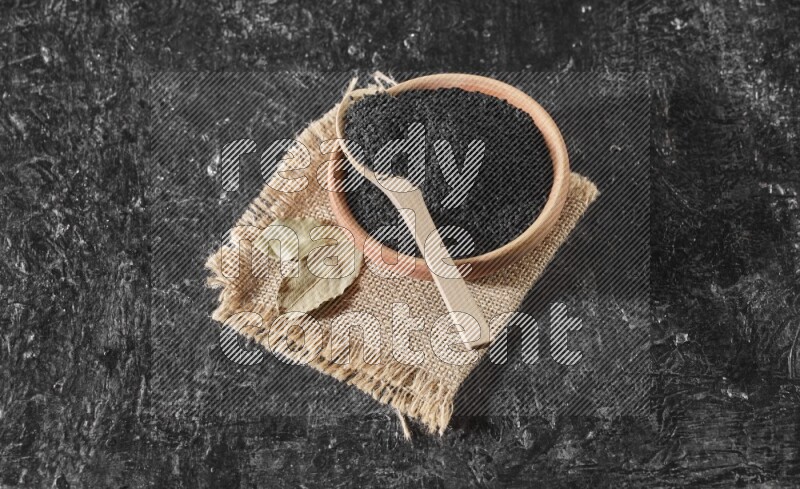 A wooden bowl full of black seeds with wooden spoon full of the seeds on it on a burlap fabric on a textured black flooring