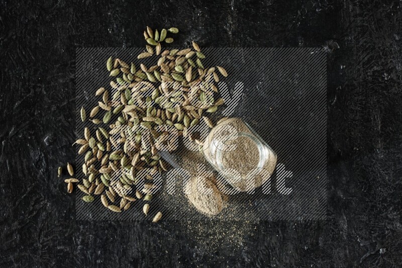 A glass spice jar and a metal spoon full of cardamom powder and cardamom seeds spreaded on textured black flooring