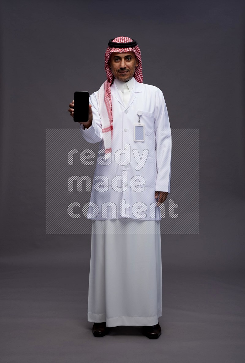 Saudi man wearing thob with lab coat and shomag with pocket employee badge standing showing phone to camera on gray background