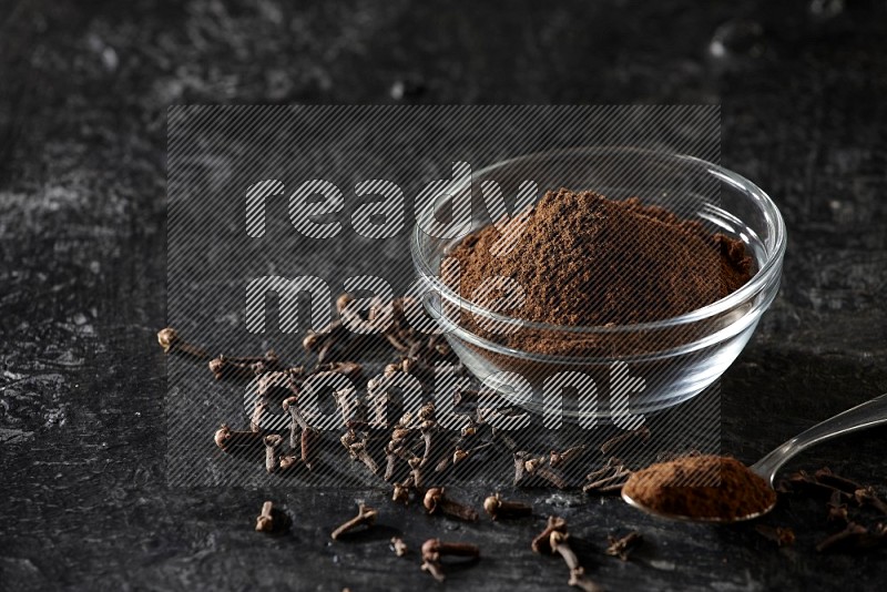 A glass bowl and a metal spoon full of cloves powder with gloves grains beside them on a textured black flooring