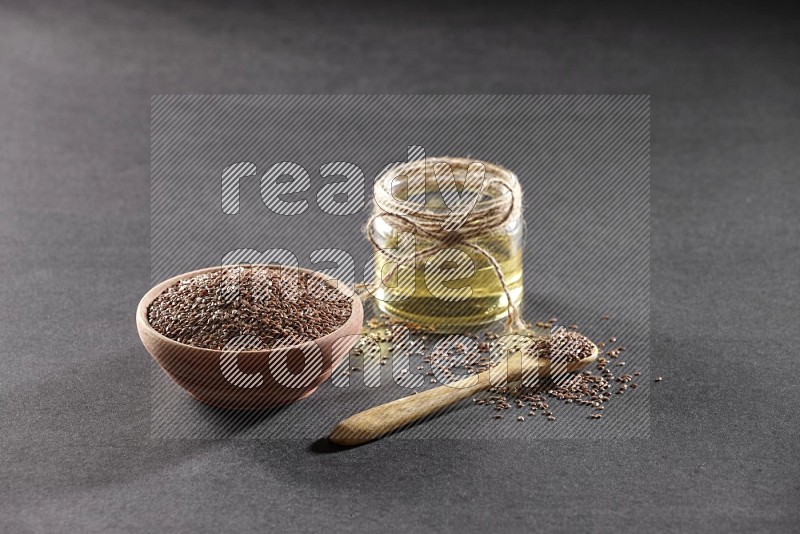 A wooden bowl and wooden spoon full of flaxseeds with a glass jar of flaxseeds oil on a black flooring