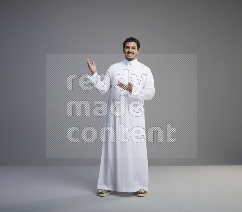 A Saudi man standing wearing thob with face painting interacting with the camera on gray background