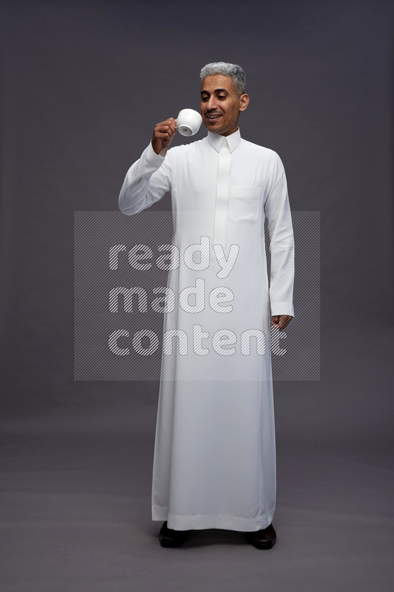 Saudi man wearing thob standing holding mug on gray background