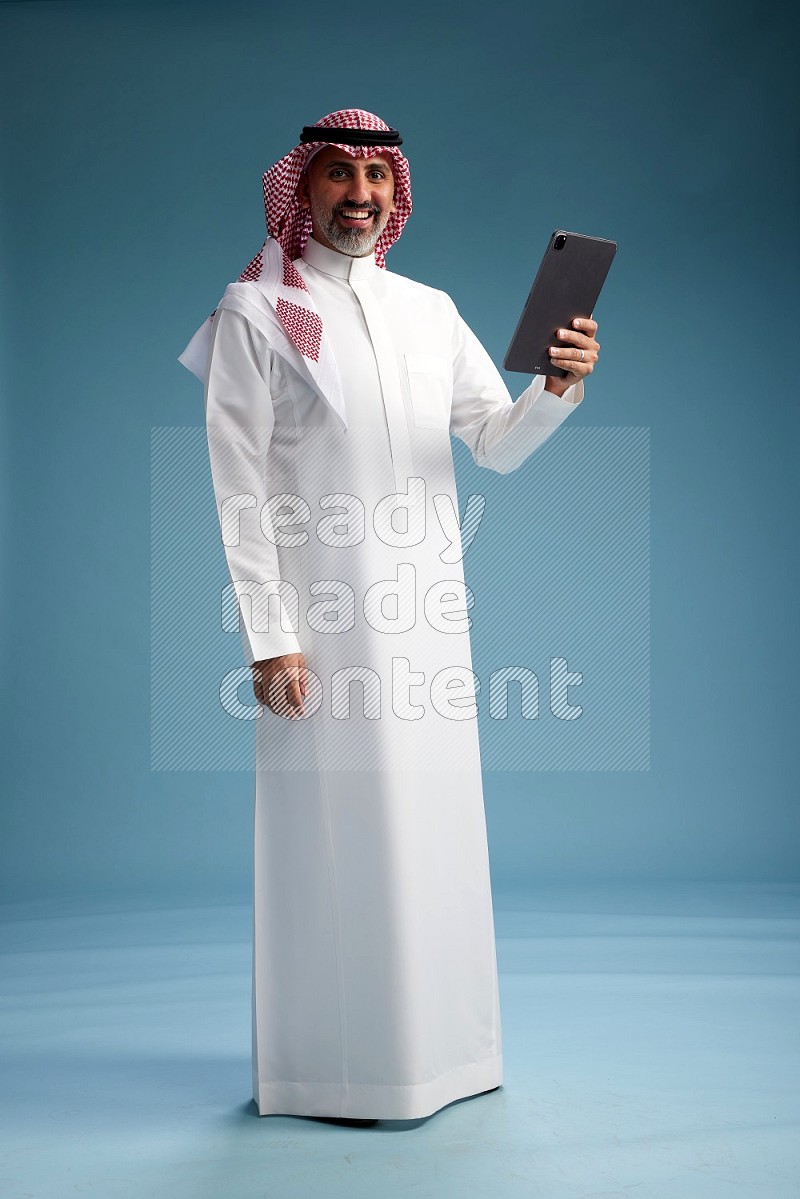 Saudi man wearing thob and shomagh with tablet on Blue background