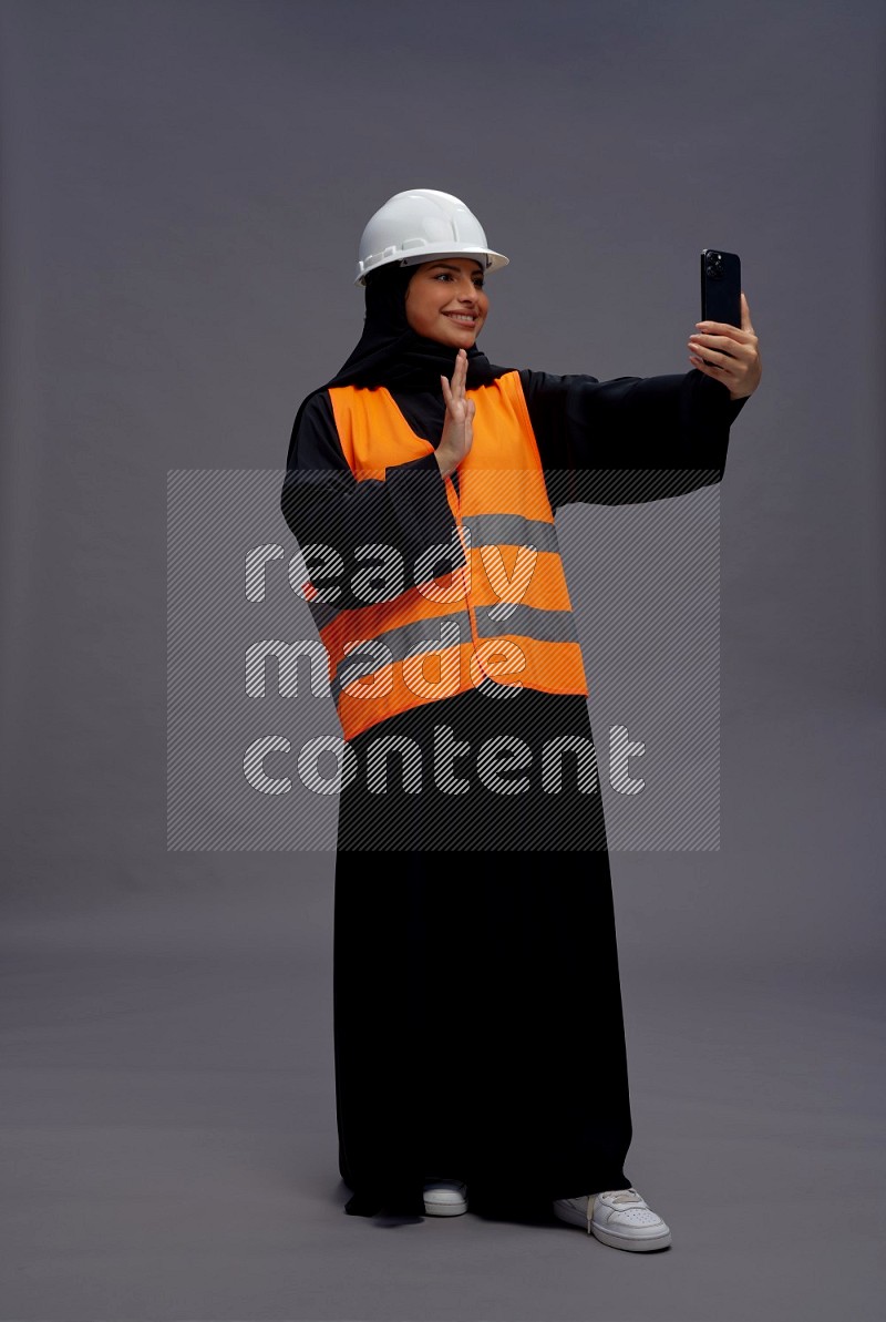 Saudi woman wearing Abaya with engineer vest standing taking selfie on gray background