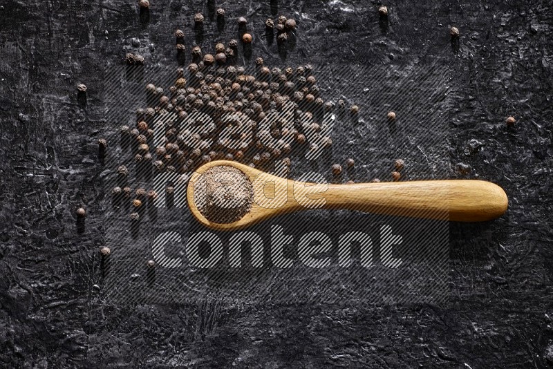 حبوب الفلفل الأسود في ملعقة خشبية مع حبوب الفلفل منتشرة علي خلفية سوداء