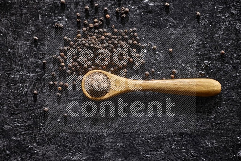 A wooden spoon full of black pepper powder and black pepper beads spread on a textured black flooring