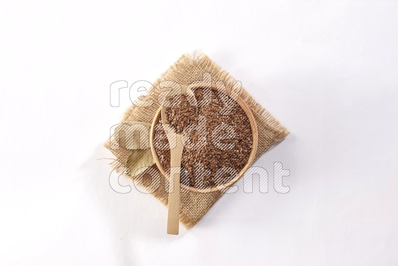 وعاء خشبي ممتلئ بحبوب بذر الكتان مع ملعقة خشبية ممتلئة علي قطعة من القماش علي خلفية بيضاء