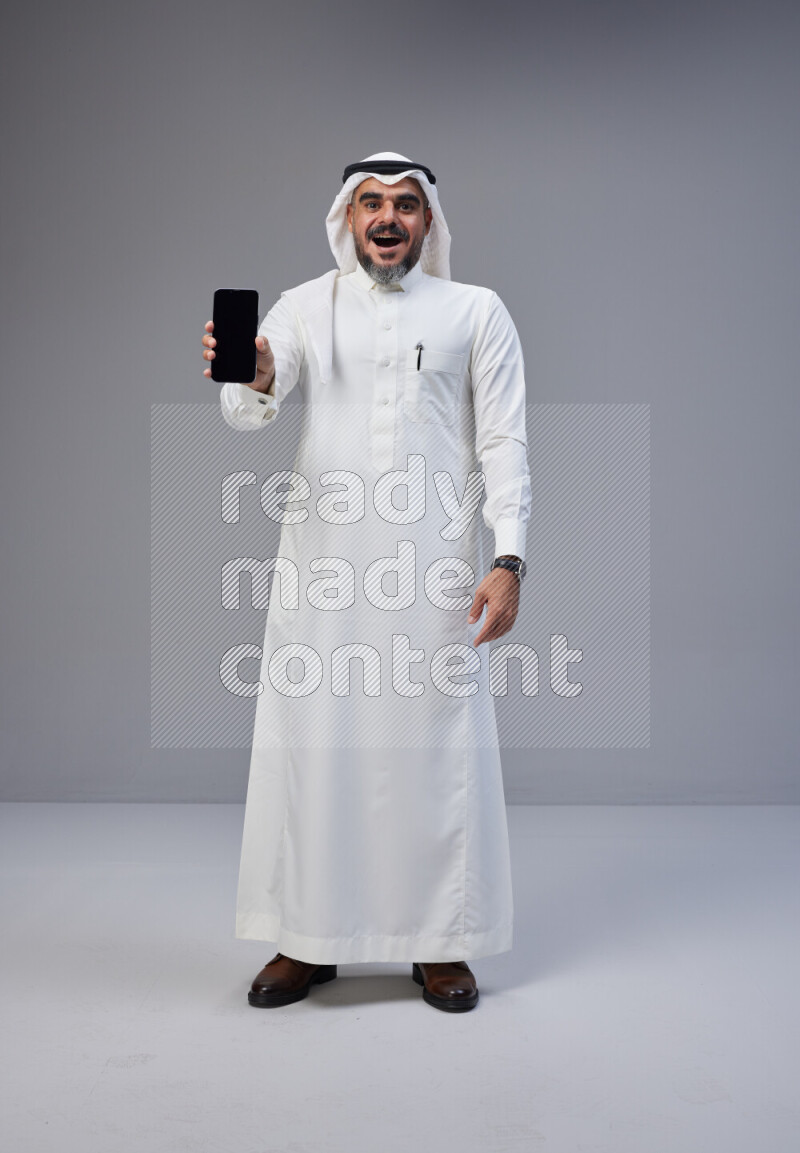 Saudi man Wearing Thob and white Shomag standing showing phone to camera on Gray background