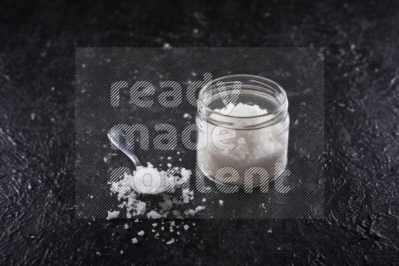 A glass jar full of coarse sea salt crystals on black background