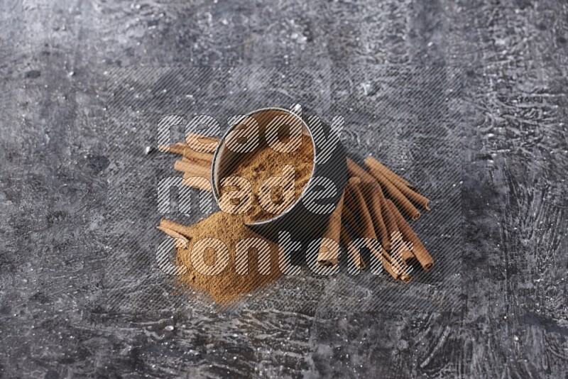 Black pottery bowl over filled with cinnamon powder and cinnamon sticks around the bowl on a textured black background