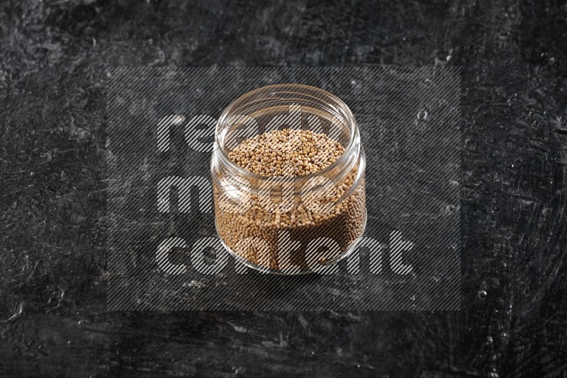 A glass jar full of mustard seeds on a textured black flooring in different angles