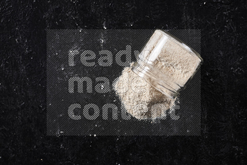 A glass jar full of onion powder flipped with some spilling powder on black background