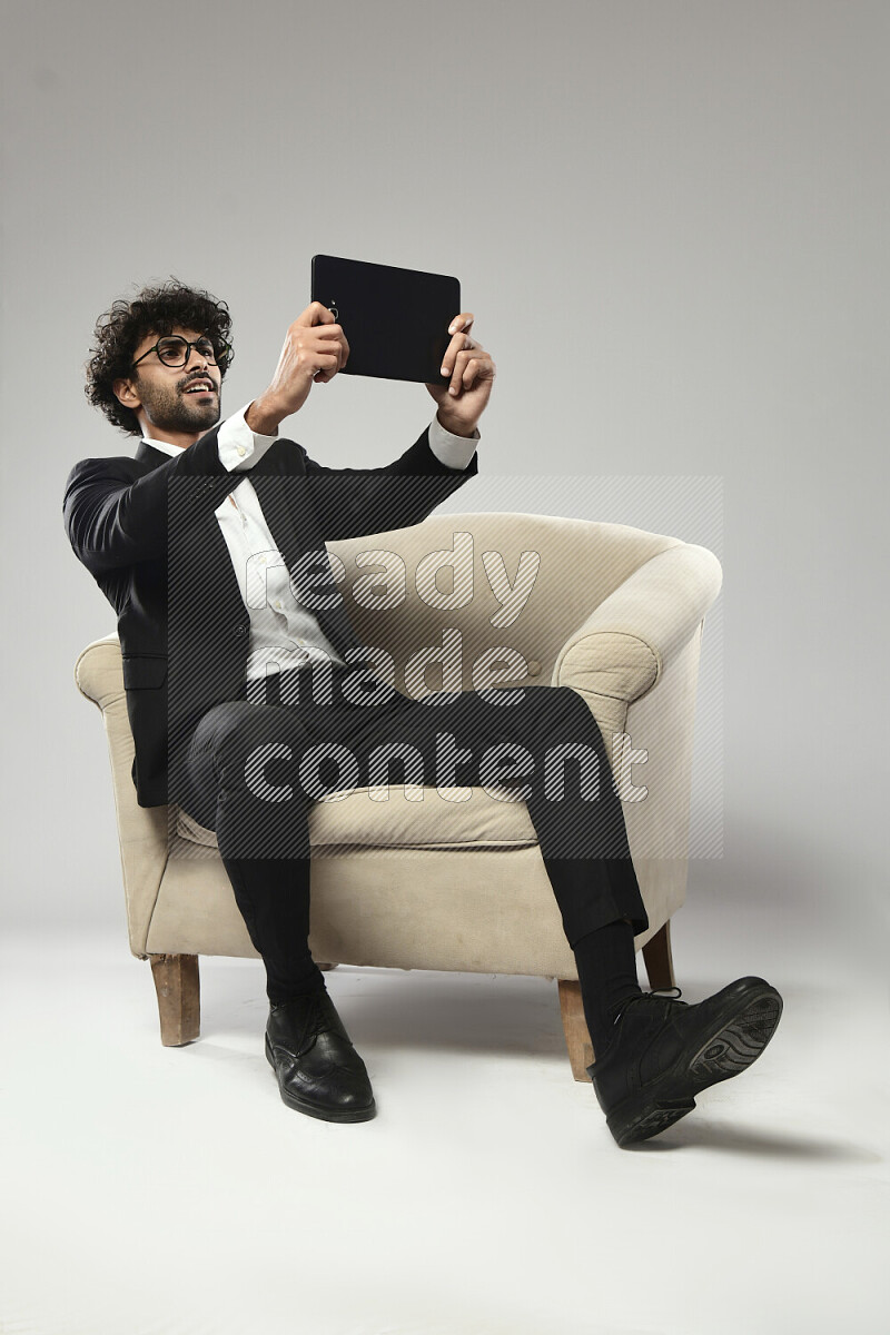 رجل يرتدي ملابس رسمية يجلس علي كرسي و يلعب علي تابلت علي خلفية بيضاء