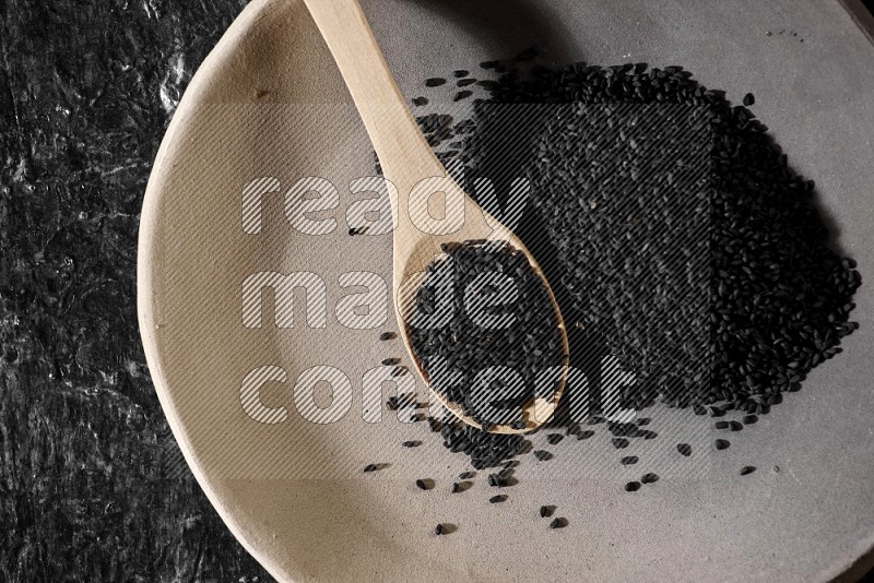 A multicolored pottery plate full of black seeds and wooden spoon full of seeds on a textured black flooring
