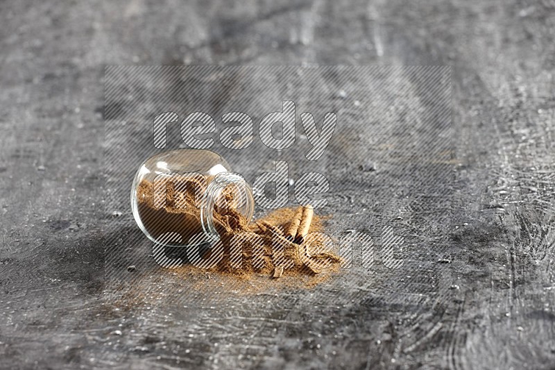 Flipped glass herbs jar full of cinnamon powder with cracked cinnamon sticks on a textured black background