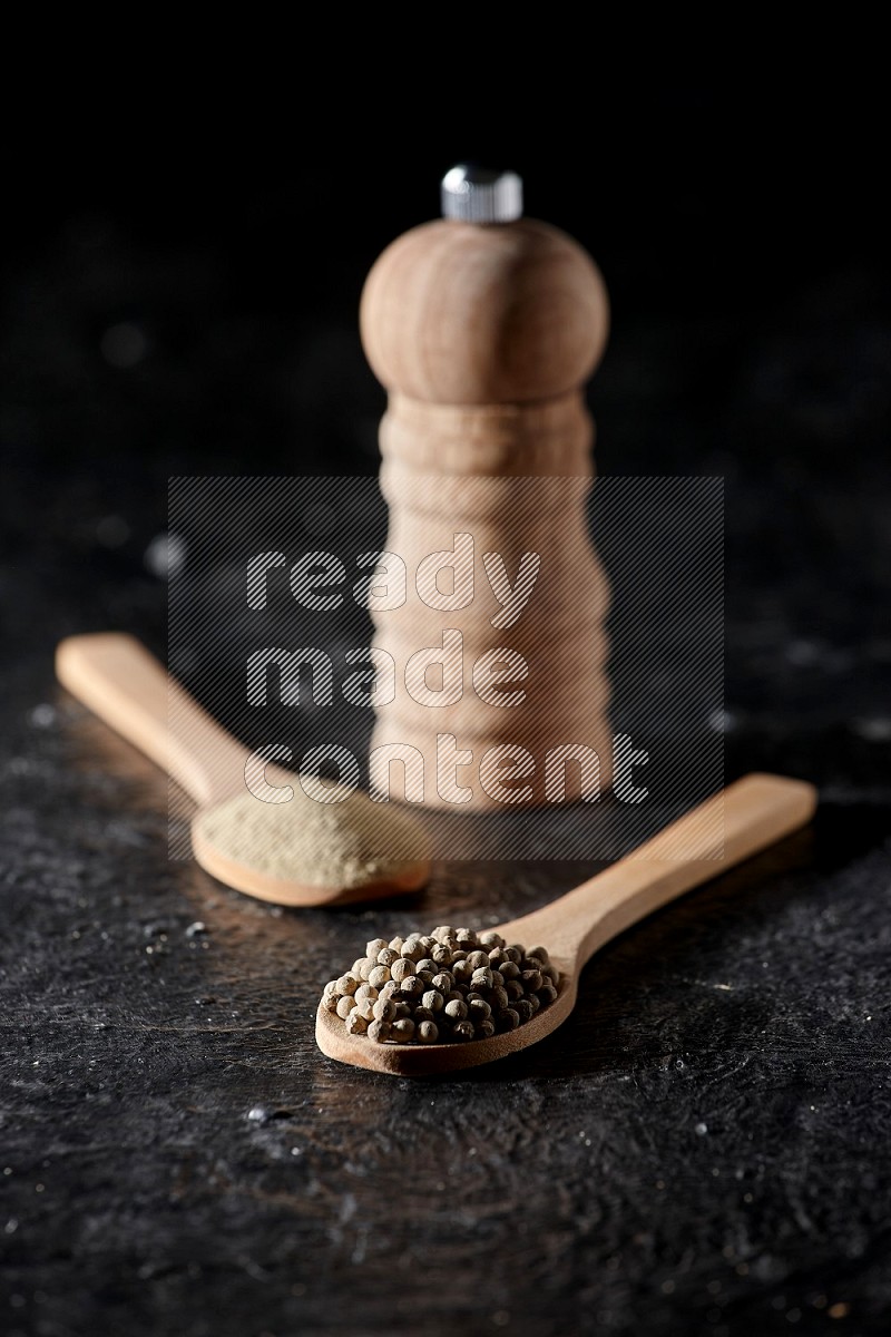 2 wooden spoons one full of white pepper powder and the other with pepper beads and a wooden pepper mill on textured black flooring