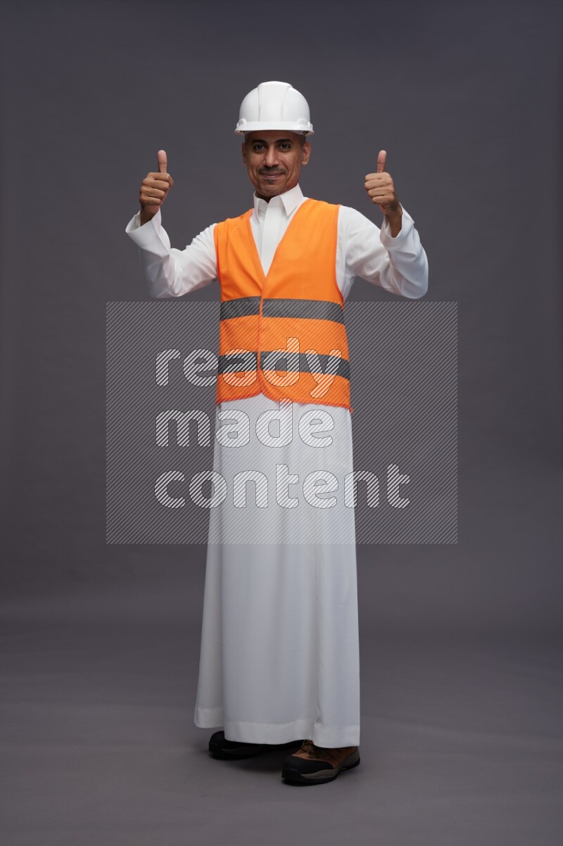 Saudi man wearing thob with engineer vest standing interacting with the camera on gray background