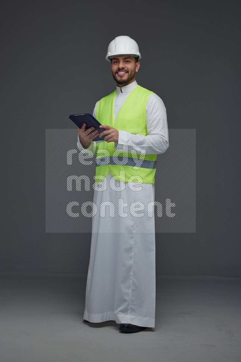 A Saudi man wearing Thobe with a yellow safety vest and white helmet standing and using his tablet different angles eye level on a gray background