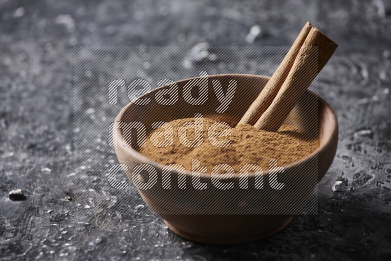 Wooden bowl full of cinnamon powder and a cinnamon stick on a textured black background