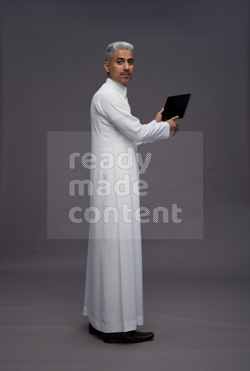 Saudi man wearing thob standing working on tablet on gray background