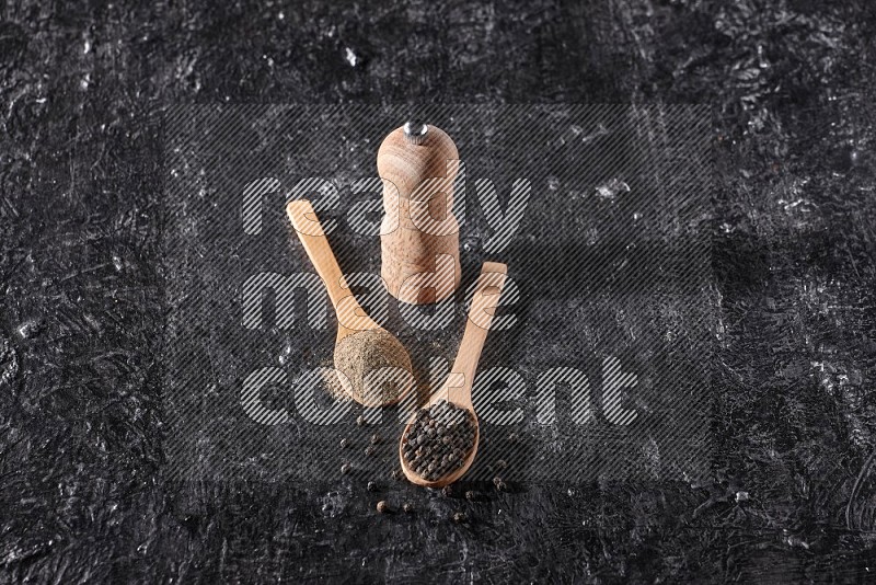بودرة وحبوب الفلفل الأسود في ملعقتان خشبيتان مع مطحنة خشبية علي خلفية سوداء