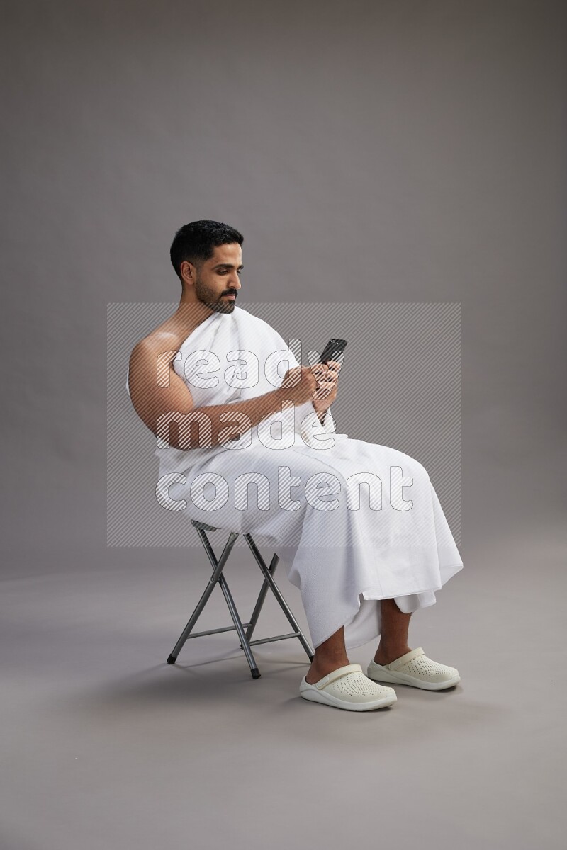 A man wearing Ehram sitting on chair texting on phone on gray background
