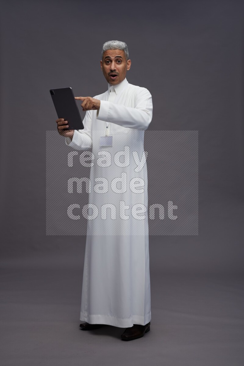 Saudi man wearing thob with neck strap employee badge standing working on tablet on gray background