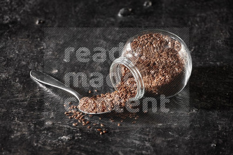 وعاء زجاجي مقلوب ممتلئ بحبوب بذر الكتان والحبوب متناثرة منه مع ملعقة معدنية ممتلئة علي خلفية سوداء