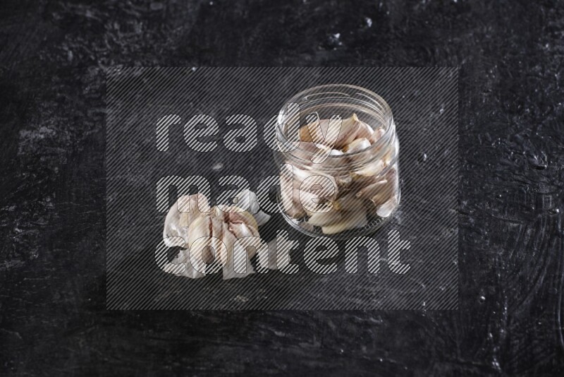 A glass jar full of garlic cloves with a whole garlic bulb beside it on a textured black flooring