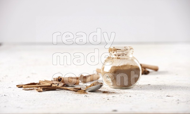 Herbal glass jar full cinnamon powder and a metal spoon surrounded by cinnamon sticks on a white background