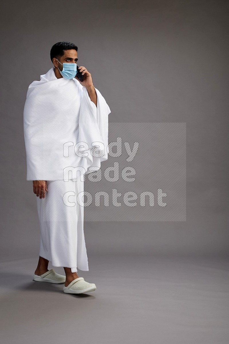 A man wearing Ehram with face mask Standing talking on phone on gray background