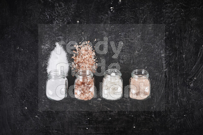 4 glass jars filled with table salt, coarse sea salt, fine himalayan salt and coarse himalayan salt on black background