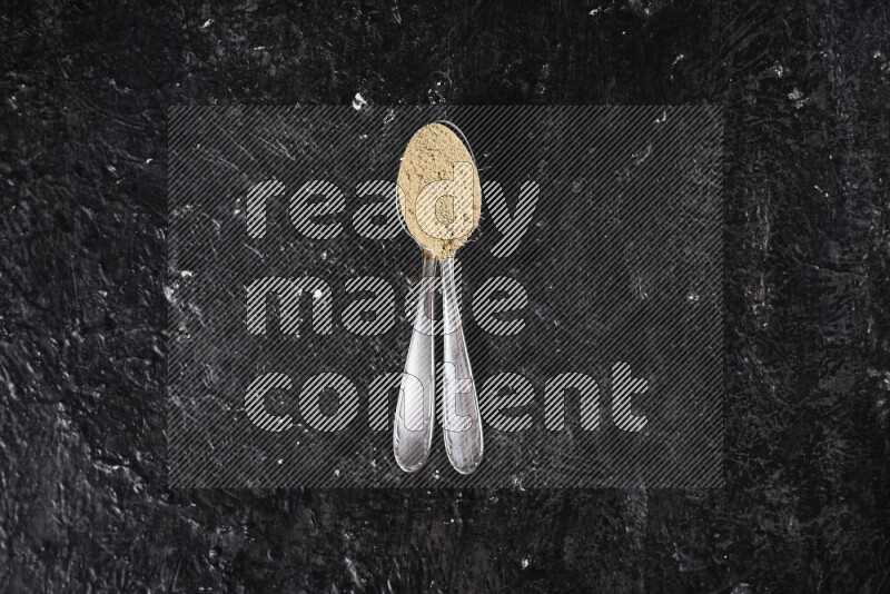 Two metal spoons full of ground ginger powder on black background