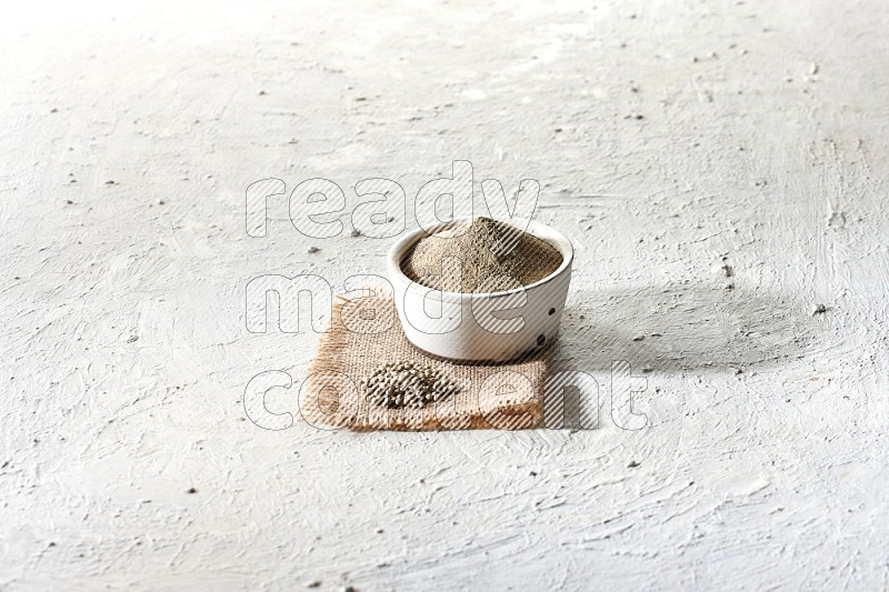 وعاء فخاري أبيض ممتلئ ببودرة الفلفل الأبيض موضوع على قطعة من الخيش مع حبوب الفلفل ومطحنة خشبية على أرضية بيضاء