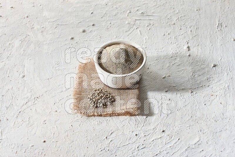 White pottery bowl full of white pepper powder set on a burlap piece of fabric with pepper beads and wooden pepper grinder on textured white flooring