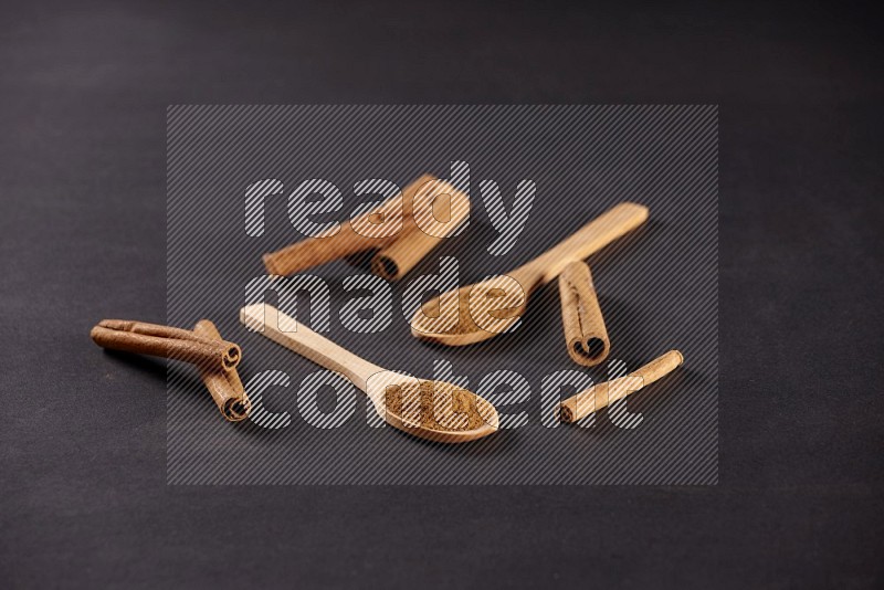 Two wooden spoons full of cinnamon powder with cinnamon sticks on black background