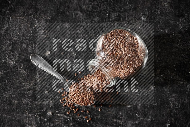 A glass spice jar full of flaxseeds flipped and seeds spread out with a metal spoon full of the seeds on a textured black flooring