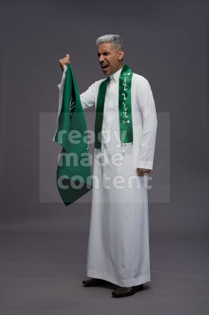 Saudi man wearing thob standing holding Saudi flag on gray background