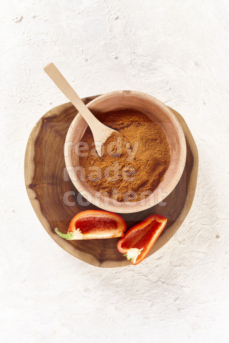 A wooden bowl full of ground paprika powder and sliced red bell pepper beside it all on a wooden tray on white background