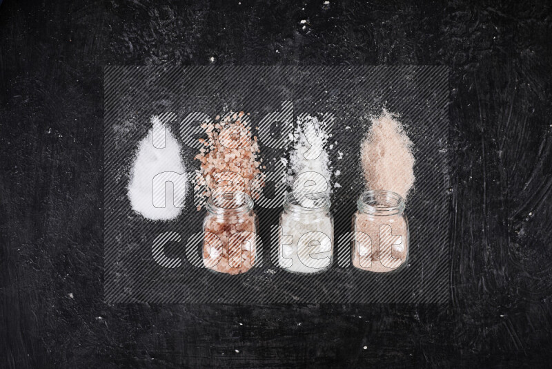 4 glass jars filled with table salt, coarse sea salt, fine himalayan salt and coarse himalayan salt on black background