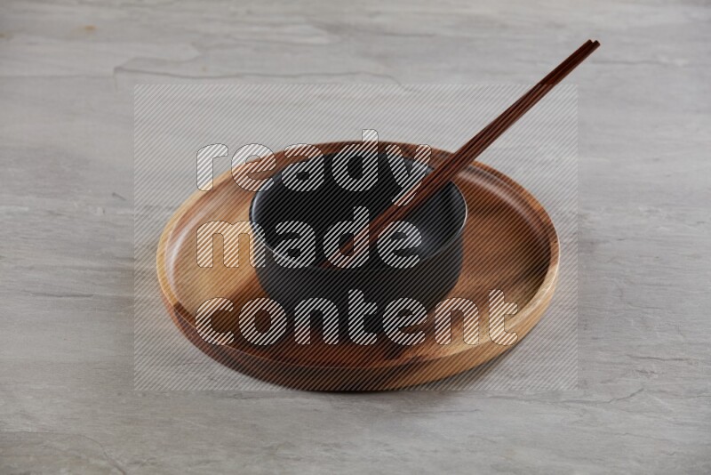 black ceramic round bowl on top of brown wood round plate and wood chopsticks, on grey textured countertop