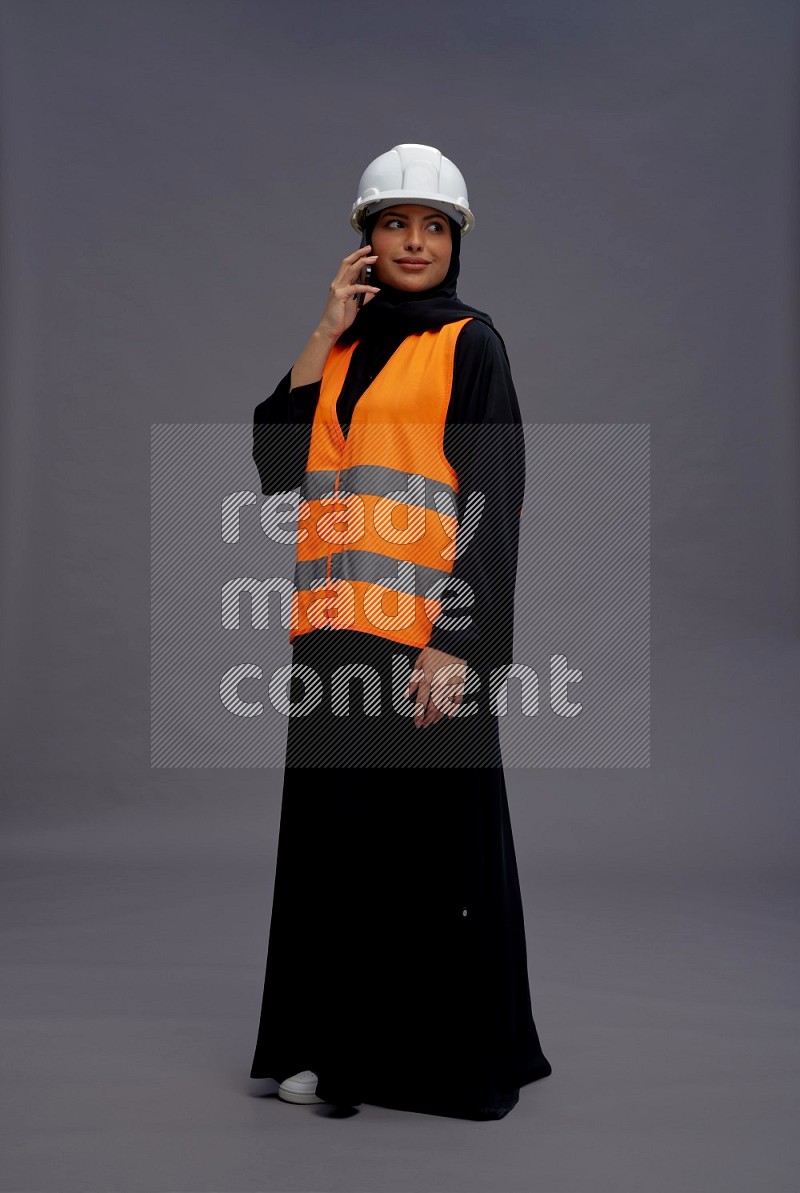 Saudi woman wearing Abaya with engineer vest standing talking on phone on gray background
