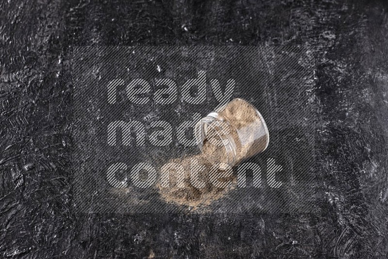 A flipped glass jar full of black pepper powder on a textured black flooring