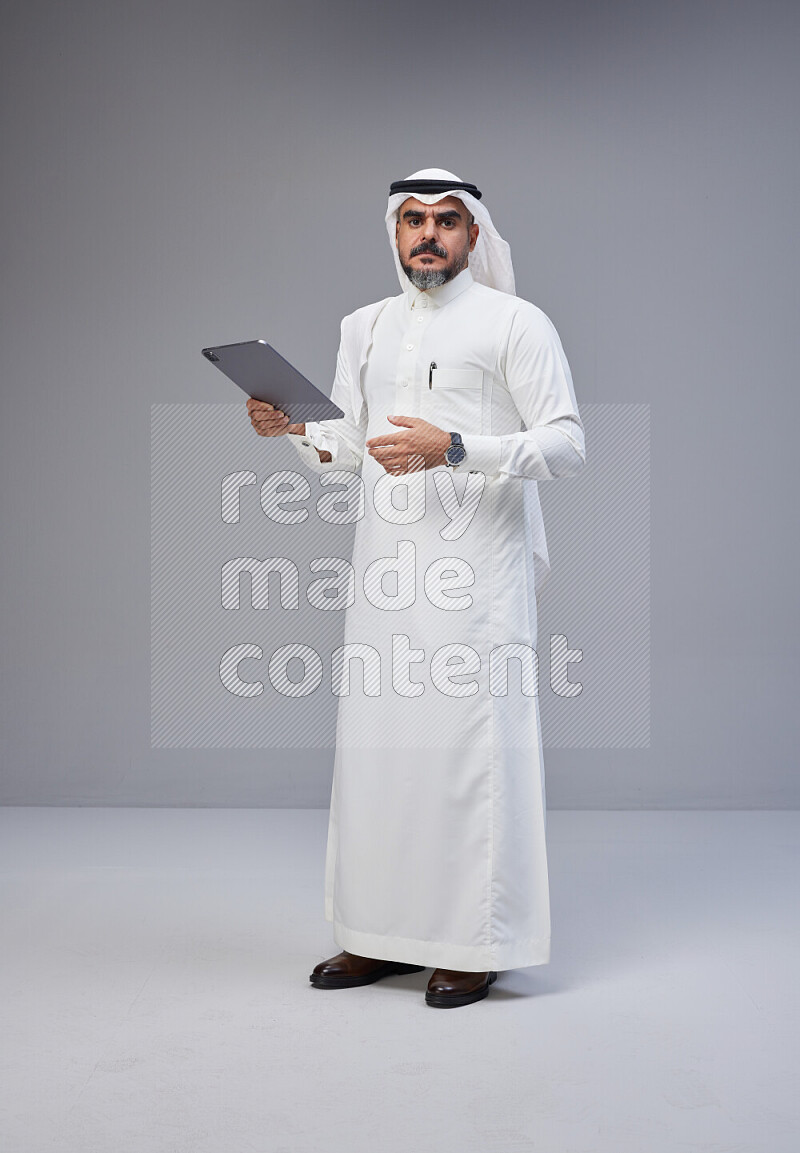 Saudi man Wearing Thob and white Shomag standing working on tablet on Gray background