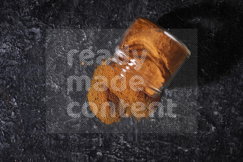 A glass jar full of ground paprika powder flipped with some spilling powder on black background