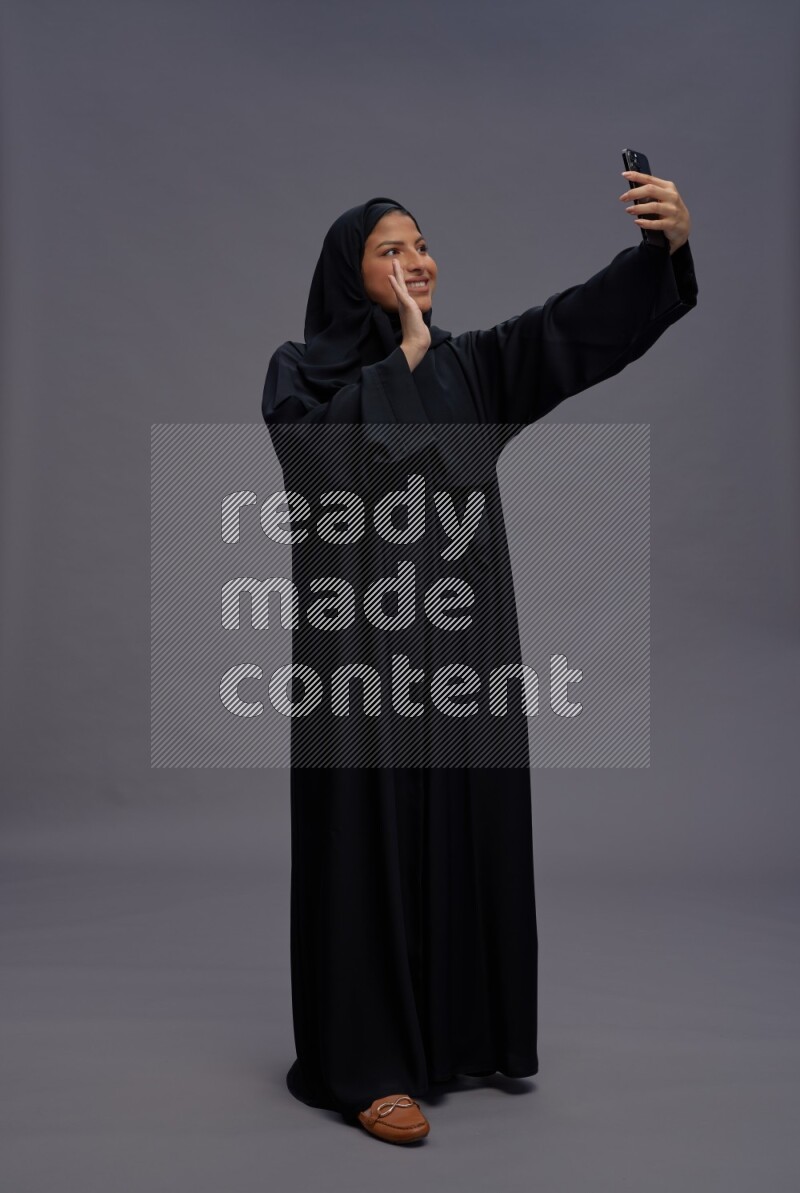 Saudi woman wearing Abaya standing taking selfie on gray background